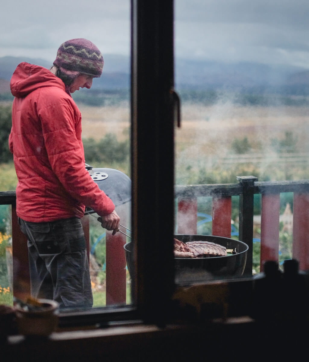 Farmer cooking local Lynbreck meat on BBQ viewed through the window of a cabin with Cairngorms hills in the background.